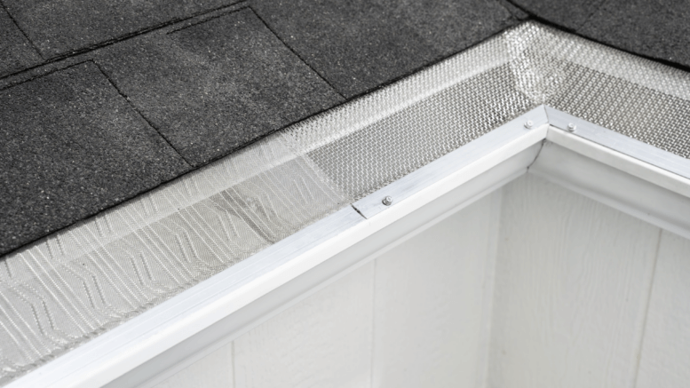 Close-up view of a house roof corner featuring dark asphalt shingles and a white rain gutter with Gutterglove mesh guard, utilizing V-Bend Technology to prevent debris from entering the gutter.